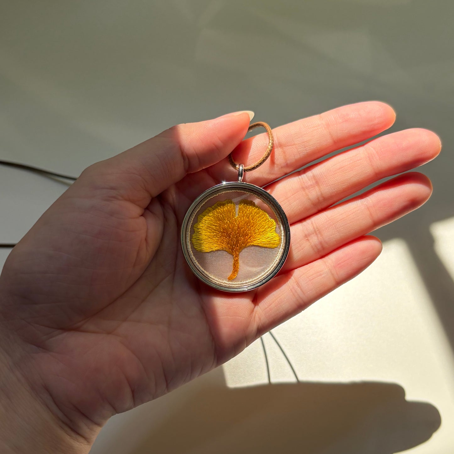 An embroidered ginkgo leaf pendant held in the sunlight.